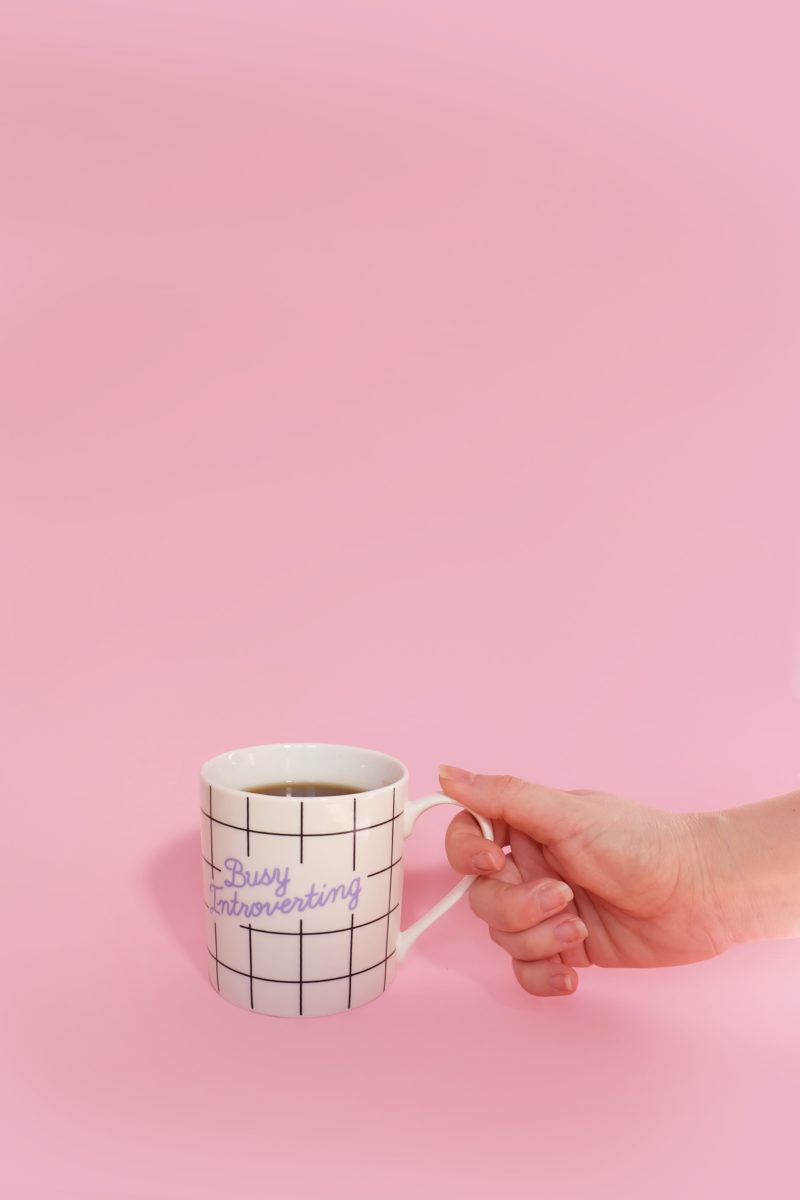 Coffee mug with words "Busy Introverting" against a pink background, held with a hand